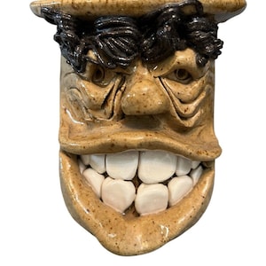 May include: A ceramic face-shaped mug with a wide, toothy grin. The mug is tan with dark brown hair and eyebrows. The teeth are white, and the eyes are small and dark. The mug has a unique, expressive design.
