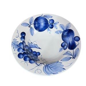 May include: White ceramic serving bowl with a blue fruit and leaf design. The bowl features a central well and is decorated with hand-painted blue grapes, apples, and leaves. The rim is slightly curved, and the overall design is elegant.