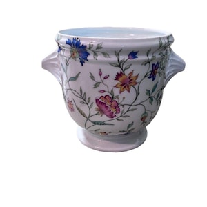 May include: White ceramic vase with a floral pattern featuring pink, blue, and yellow flowers. The vase has two handles and a pedestal base. The floral design is set against a white background.