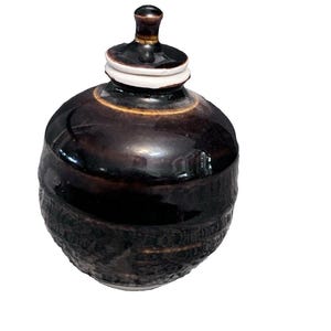 May include: A dark brown ceramic jar with a lid. The jar has a rounded body with a textured lower section and a smooth, glossy upper section. The lid is topped with a small knob and a white band.