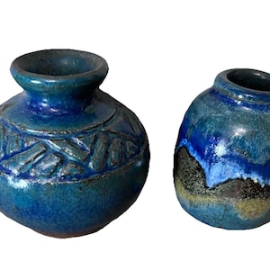 May include: Two handcrafted ceramic vases in shades of blue. The larger vase has a textured, carved design around its body. The smaller vase has a glossy, layered appearance with dark and light blue hues. Both have dark openings.
