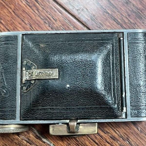 May include: A vintage black leather camera with a silver metal frame. The camera features a textured leather surface and a small metal plate with the word "Voigtlander". The camera is closed and has a metal clasp.