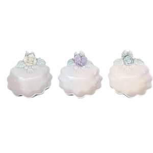 May include: Three ceramic trinket dishes in a row, each with a scalloped edge and topped with a floral arrangement. The flowers are in shades of white, purple, and blue, with green leaves. The dishes are a soft, pearly white.