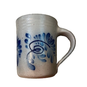 May include: A cylindrical ceramic mug with a speckled off-white glaze and a single handle. The exterior features hand-painted blue floral motifs with swirling leaf patterns arranged vertically around the body of the vessel.