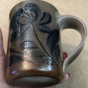 May include: A cylindrical ceramic mug with a textured, mottled brown and tan glaze. The exterior features a dark blue, hand-painted floral design with a prominent central flower and leaves. It has a sturdy, curved handle on the right side.