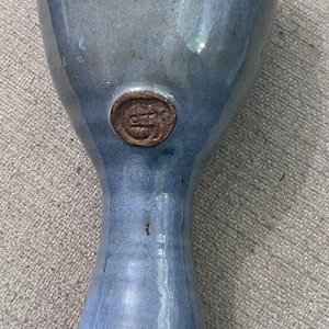 May include: A handmade, blue ceramic vase with a flared base and a wide rim. The vase has a smooth, glossy finish and a small, circular emblem near the top. The vase is approximately 8 inches tall.