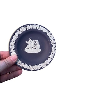 May include: A small, round, dark blue decorative plate with a white relief design. The plate features a central scene of figures and a border of white floral patterns. The plate is held by a hand.