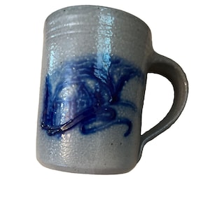 May include: A cylindrical ceramic mug with a speckled light gray finish and a textured surface. The mug features a prominent, hand-painted cobalt blue abstract design on the front and includes a sturdy, curved handle on the right side.