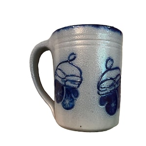 May include: A light gray ceramic mug with a textured surface and a dark blue rim. The mug features two embossed blue designs of winter mittens, each decorated with a snowflake pattern. It has a curved handle on the left side and a cylindrical body.