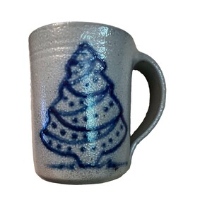 May include: A light gray speckled ceramic mug with a textured surface and a single handle. The front of the mug features a hand-painted dark blue graphic of a stylized Christmas tree with dotted details, centered on the cylindrical body.