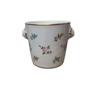 May include: A white porcelain bucket with a gold rim and base, featuring hand-painted floral motifs in pink, blue, and orange. The vessel includes two side handles shaped like sculpted animal heads with gold accents, providing a decorative finish.