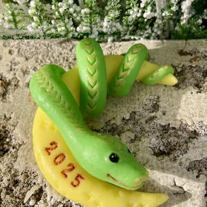 May include: A handmade sculpture of a green snake on a yellow crescent moon. The snake has gold detailing and the number "2025" is on the moon. The background features greenery with white accents.