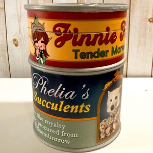 Op de afbeelding: Een cilindrische container met twee gestapelde labels. Het bovenste label is rood en geel met de tekst Finnie Tender Mors en een elfenillustratie. Het onderste label is blauw en groen met de tekst Phelia's Succulents en een witte hond.