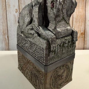 May include: A rectangular stone-textured box featuring a sculpted creature with a single red eye on the lid. The base displays intricate relief carvings of figures and circular patterns, while the lid includes etched runic-style symbols.