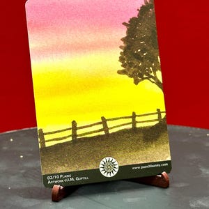 May include: A landscape art card featuring a watercolor painting of a sunset over a field with a fence and a tree. The sky transitions from pink to yellow. The card is displayed on a small wooden easel. Text on the card reads "02/10 Plains" and "Artwork ©J.M. Guptill".