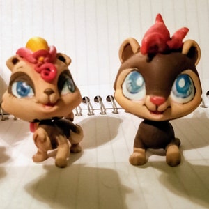 Pode incluir: Dois esquilos de brinquedo marrons e bege com olhos azuis. Um esquilo tem um moicano vermelho e amarelo e o outro tem um redemoinho rosa e amarelo na cabeça.