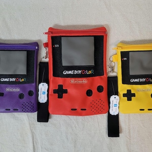 May include: Three colorful Game Boy Color-themed zipper pouches in purple, red, and yellow. Each pouch features a black screen, the words "Game Boy Color" and "Nintendo", and a black wrist strap with a silver clasp.