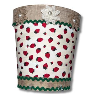 May include: A white fabric storage bin with a green scalloped trim and a brown burlap top. The bin is decorated with a white flower with a pearl center and a red strawberry pattern.