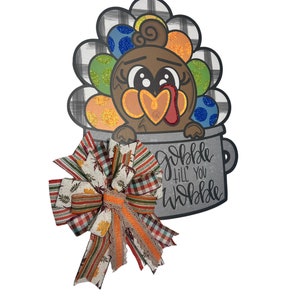 May include: A Thanksgiving-themed door hanger featuring a cartoon turkey in a pot with the text "Gobble 'til you wobble". The turkey is brown with a red wattle and has a heart-shaped body. The pot is gray with a white rim. A bow with red, orange, and white plaid ribbon is attached to the pot.