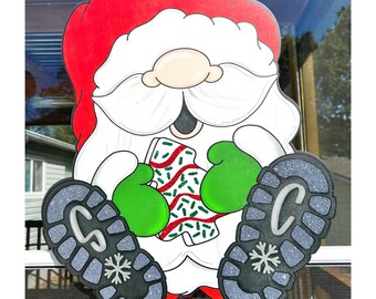 May include: A wooden Santa Claus decoration with a red hat, white beard, and green mittens. The Santa holds a Christmas tree-shaped cookie with red and green sprinkles. The boots are gray with a snowflake and the letter "C".