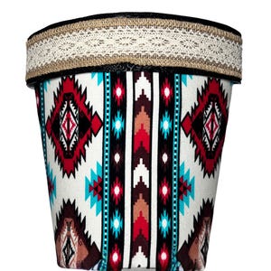 May include: A white planter with a geometric pattern in shades of red, blue, and brown. The planter has a brown and white lace trim around the top.