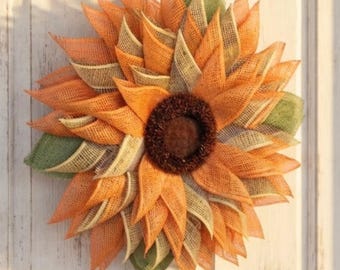 May include: A decorative wreath shaped like a sunflower, constructed from layers of orange, yellow, and green burlap fabric. The center of the wreath features a dark brown, textured circular core, mimicking the appearance of a sunflower seed head.