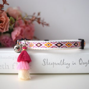May include: A white cat collar with a pink, orange, and white geometric pattern. The collar has a silver bell and a pink, light pink, and cream tassel.