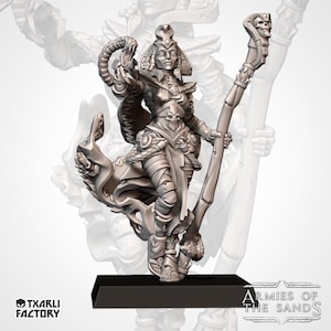 Op de afbeelding: Een grijze hars miniatuurfiguur van een persoon met een versierde hoofdtooi en gewikkelde kleding, die een staf met een schedel en in de andere hand een kronkelende slang vasthoudt, staand op een zwarte rechthoekige basis met tekst.