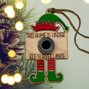 May include: Wooden Christmas ornament shaped like an elf camera with the text "THIS HOME IS UNDER ELF SURVEILLANCE." The ornament has a green elf hat, red and green legs, and a black camera lens. A brown string is attached for hanging.