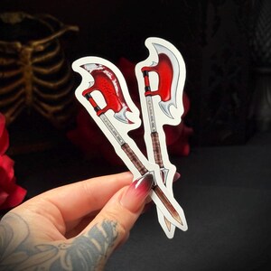 May include: Two stickers of a red and silver battle axe with a brown handle. The axe heads are a bright red colour, and the blades are silver. The stickers are held in a hand with a dark background.