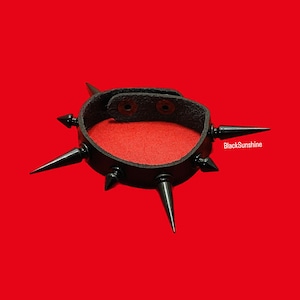 May include: Black leather bracelet with multiple sharp metal spikes. The bracelet is on a red background.