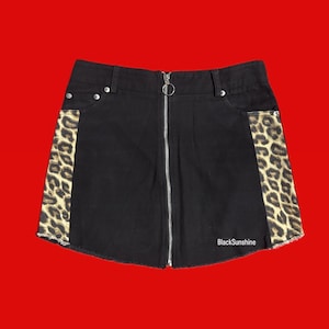 Może przedstawiać: Czarna dżinsowa mini spódniczka z zapięciem na zamek z przodu i bocznymi panelami w panterkę. Spódniczka ma surowy dół i mały metalowy pierścień na suwaku. Tekst "BlackSunshine" jest widoczny po prawej stronie u dołu.