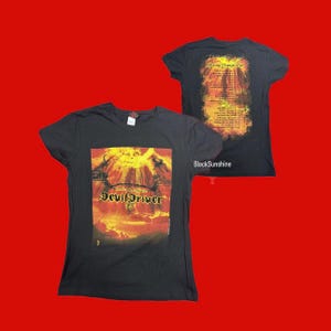 Puede incluir: Dos camisetas negras de la banda DevilDriver. La parte delantera de la camiseta muestra el nombre de la banda con un diseño gráfico ardiente. La parte trasera de la camiseta tiene una lista de giras con un diseño ardiente similar. Las camisetas se muestran sobre un fondo rojo.