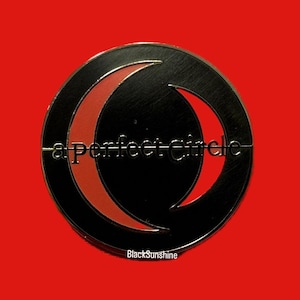 Puede incluir: Un pin circular negro y dorado con un diseño de luna creciente roja. El texto "a Perfect Circle" está escrito en blanco en el centro del pin.