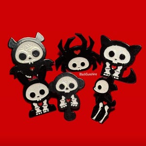 May include: A collection of black and white skeleton-themed pins in various shapes, including a bat, spider, cat, bunny, monkey, and a sitting figure. Each pin features a skull design with a red heart and is set against a red background. The text "BlackSunshine" is visible.