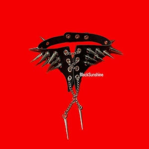 May include: Black leather choker necklace with silver-colored spikes and chain accents. The choker has a studded collar and a central design with a lace-up detail. The background is a solid red.