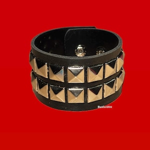May include: Black leather wristband with two rows of silver pyramid studs. The wristband has a buckle closure and is set against a red background. The text "BlackSunshine" is visible on the bottom right.