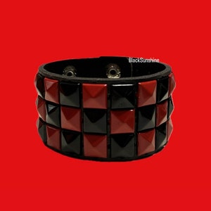 Puede incluir: Pulsera con tachuelas negras y rojas con una banda de cuero negro. La pulsera presenta filas de tachuelas cuadradas en colores negro y rojo alternados. El texto "BlackSunshine" es visible.