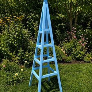 Peut inclure: Un grand obélisque de jardin en bois bleu clair en forme de pyramide à quatre faces. La structure se compose de quatre montants verticaux reliés par des traverses horizontales à intervalles réguliers, se terminant par un sommet carré.