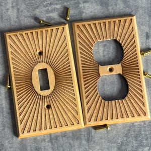 May include: Two wooden light switch covers with a sunburst design. One cover has an oval opening for a switch, the other has two holes for an outlet. Several small brass screws are scattered around the covers.