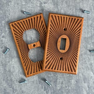 May include: Two wooden light switch covers with a sunburst design. One cover has an outlet opening, and the other has a switch opening. The covers are made of light brown wood and are accompanied by several silver screws.