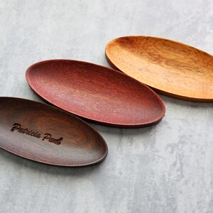 May include: Three oval-shaped wooden serving trays. The tray on the left is dark brown with the text "Patricia Peel" engraved on it. The middle tray is a reddish brown color. The tray on the right is a light brown color.