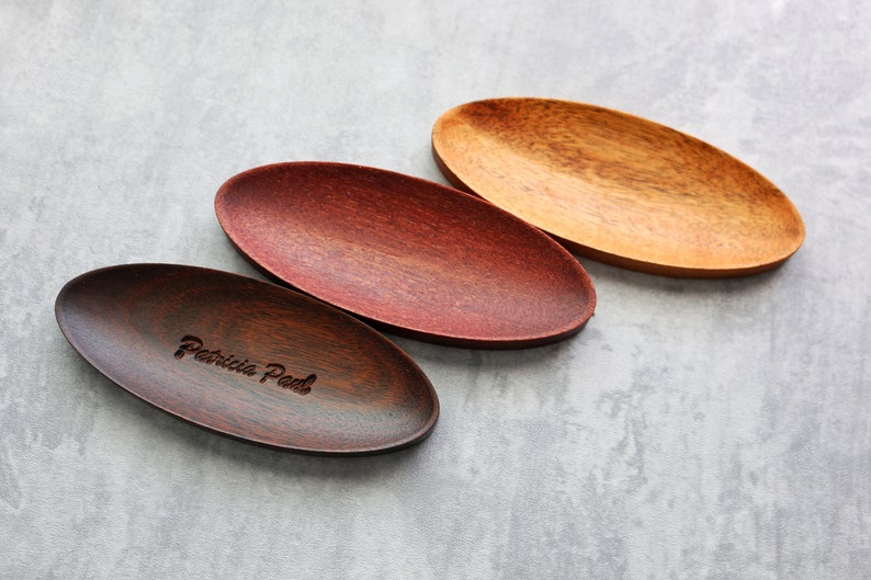 May include: Three oval-shaped wooden serving trays. The tray on the left is dark brown with the text "Patricia Peel" engraved on it. The middle tray is a reddish brown color. The tray on the right is a light brown color.