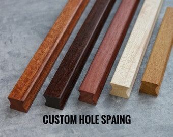 Op de afbeelding: Vijf houten kastgrepen in verschillende houttinten, waaronder bruin, rood en licht hout. De grepen zijn allemaal rechthoekig met een afgeronde rand en een uitsparing in het midden. De tekst "CUSTOM HOLE SPACING" is onder de grepen gedrukt.