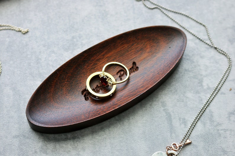 May include: A dark brown wooden oval dish with the words "Retro Park" engraved on the surface. Two gold rings with a red gemstone are resting on the dish.