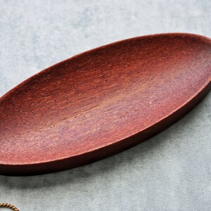 May include: A small, oval-shaped wooden dish with a reddish-brown finish. The dish is made of wood and has a smooth, polished surface.