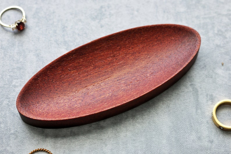 May include: A small, oval-shaped wooden dish with a reddish-brown finish. The dish is made of wood and has a smooth, polished surface.