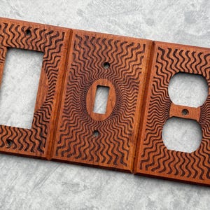 May include: Three wooden wall plate covers with a wavy pattern. The left cover has a single light switch opening, the middle cover has a single light switch opening, and the right cover has a double outlet opening.