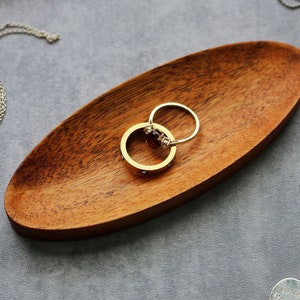May include: Two gold rings with a red gemstone on a wooden ring dish. The rings are on a gray surface.