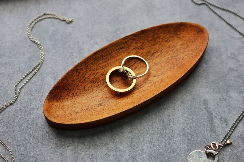 May include: Two gold rings with a red gemstone on a wooden ring dish. The rings are on a gray surface.